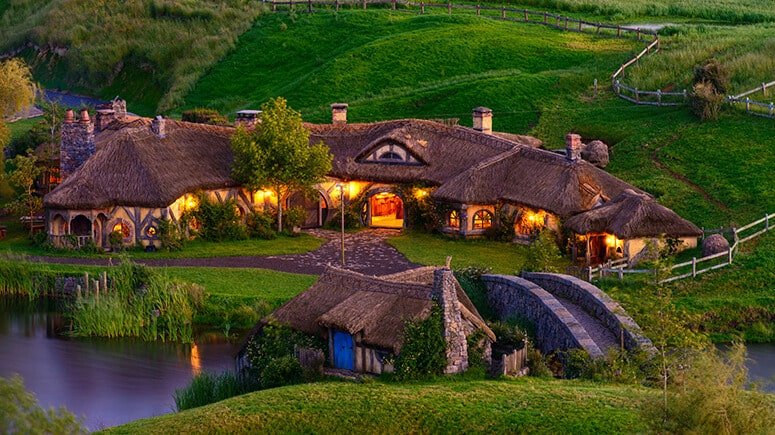 Arched stone bridge at Hobbiton with lit-up hobbit homes at twilight