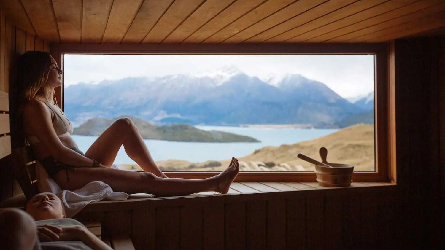 Person relaxing in a sauna with a panoramic view of lake, mountains, and natural landscape at Aroha Wellness Retreat.