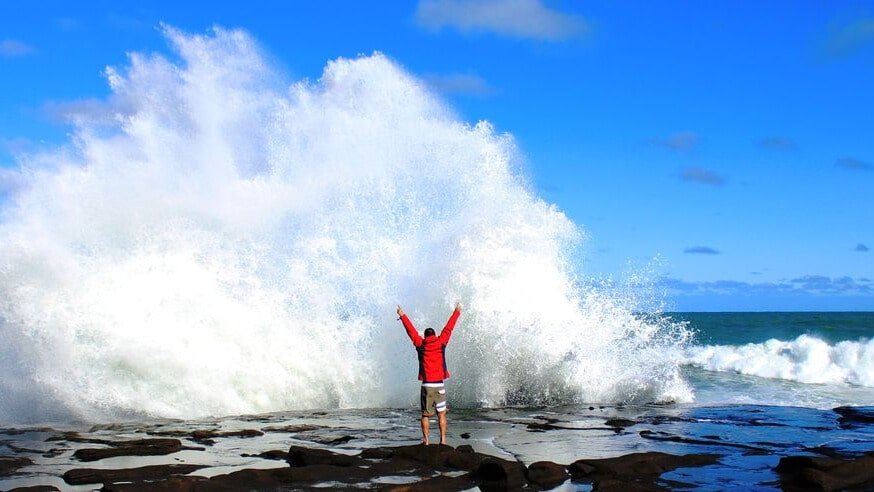 Man standing on rocky shore with ocean waves crashing and sea spray rising into the air