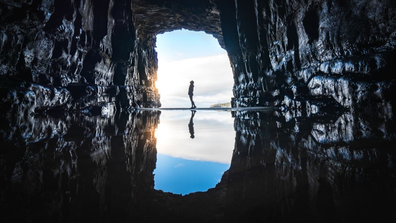 A person silhouetted at the entrance of a cave with still water reflecting the sky and cave walls