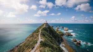 A lighthouse perched on a grassy clifftop with a winding walkway and rocky bay below
