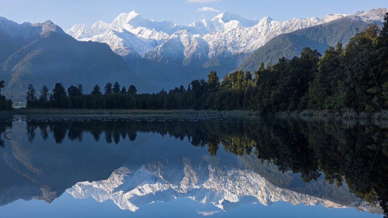 Crystal-clear lake reflecting snow-covered mountains and icy peaks on New Zealand’s West Coast.