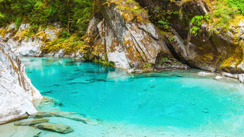 Crystal-clear blue lagoon surrounded by lush greenery and mountains on New Zealand’s West Coast.