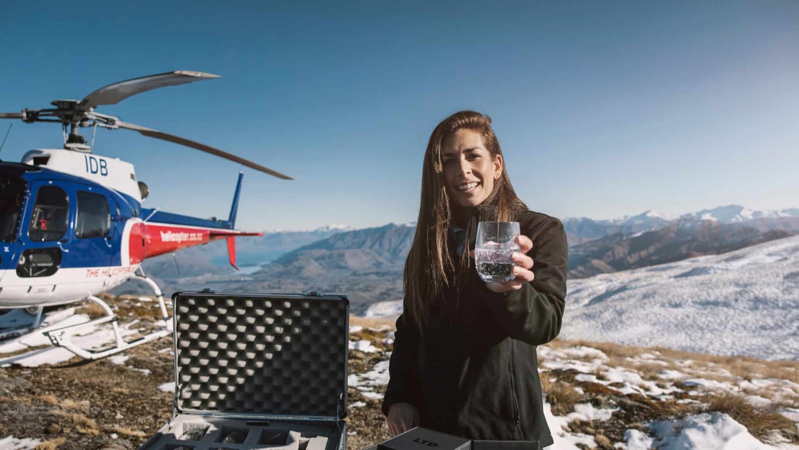 Woman holding up a gin beside a helicopter on a mountaintop during Queenstown heli-gin tour