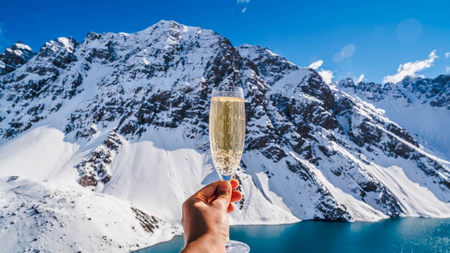 Person holding a glass of sparkling wine in front of snow-covered mountains and an ice blue glacier lake during Queenstown heli-wine tour