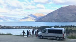 Tour van parked beside lake at Hensman Road Lookout in Queenstown, overlooking mountains and water.
