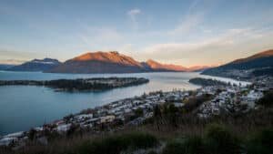 Homes along the edge of a lake with a small island and surrounding mountains in Queenstown, New Zealand.