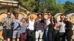 A small group holding wine glasses, standing in front of stacked wine barrels during a Queenstown wine tour.