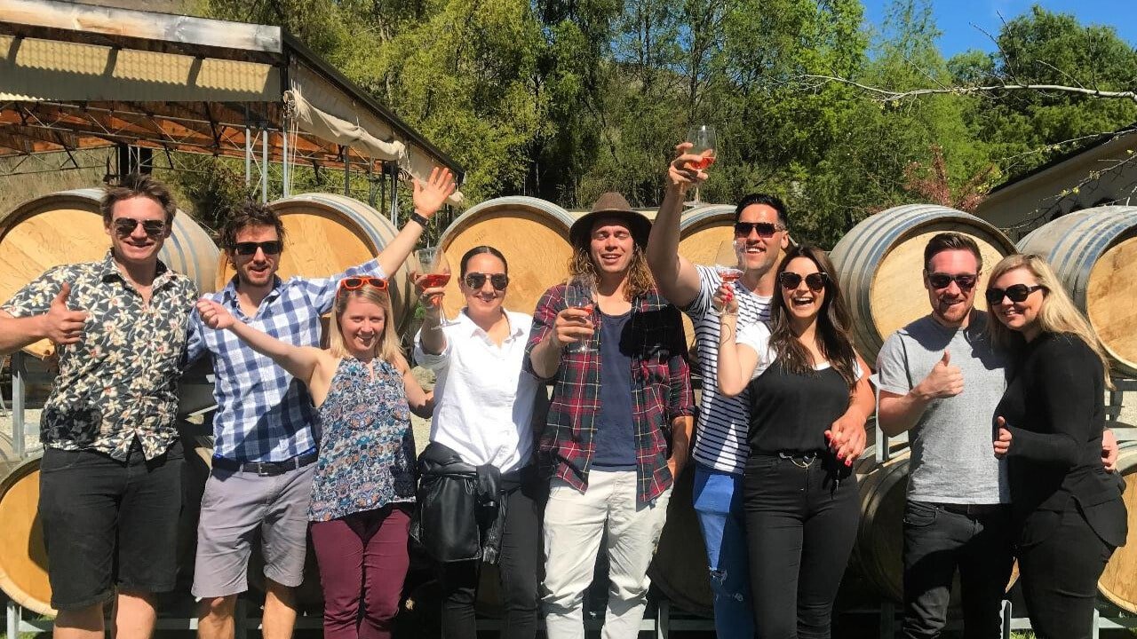 A small group holding wine glasses, standing in front of stacked wine barrels during a Queenstown wine tour.
