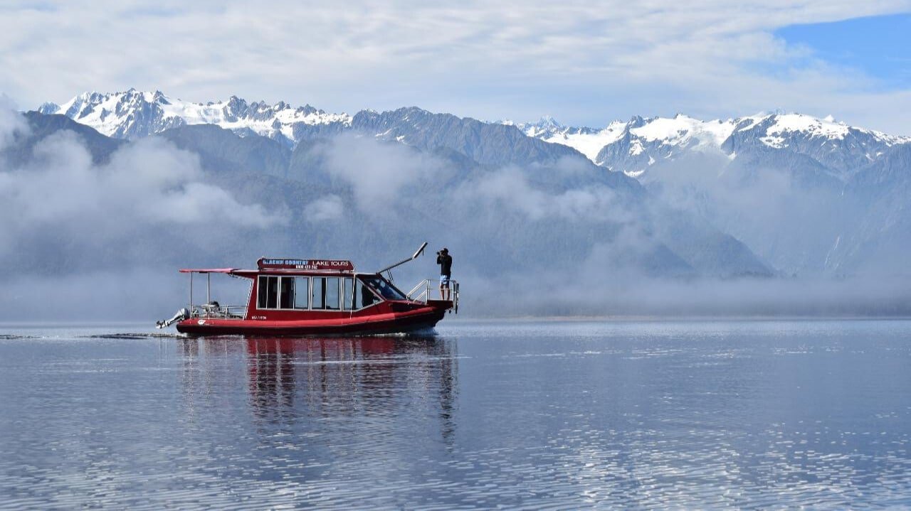 Boat cruising through mist on a fog-covered lake with mountains in the background in Franz Josef.
