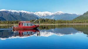 Boat cruising on a still, reflective lake with full reflections of the boat, trees, and mountains in Franz Josef.