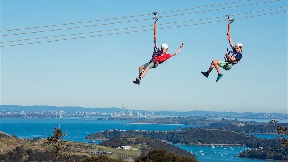 Children ziplining side-by-side with a biplane style setup, Auckland City skyline visible in the background
