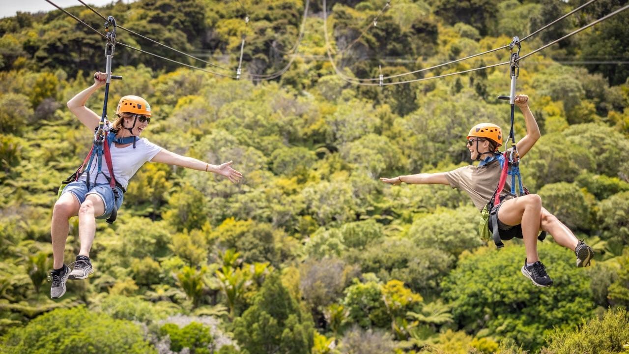 Two people ziplining side-by-side with arms reaching out towards each other on Waiheke Island