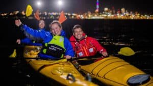 Tandem and single kayaks side by side waving with Auckland city lights glowing in the background