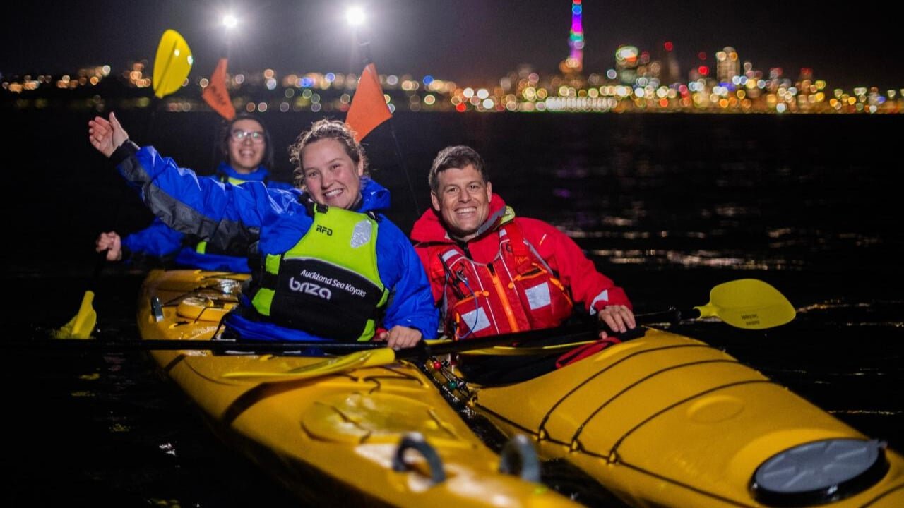 Tandem and single kayaks side by side waving with Auckland city lights glowing in the background