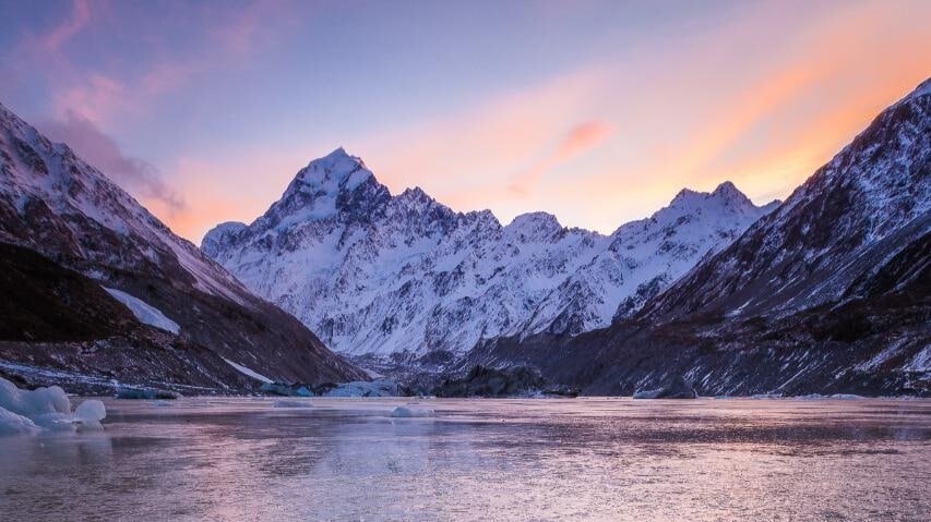 Icy lake surrounded by snow-capped mountains under an orange sunset sky on Mount Cook day tour from Christchurch via Tekapo